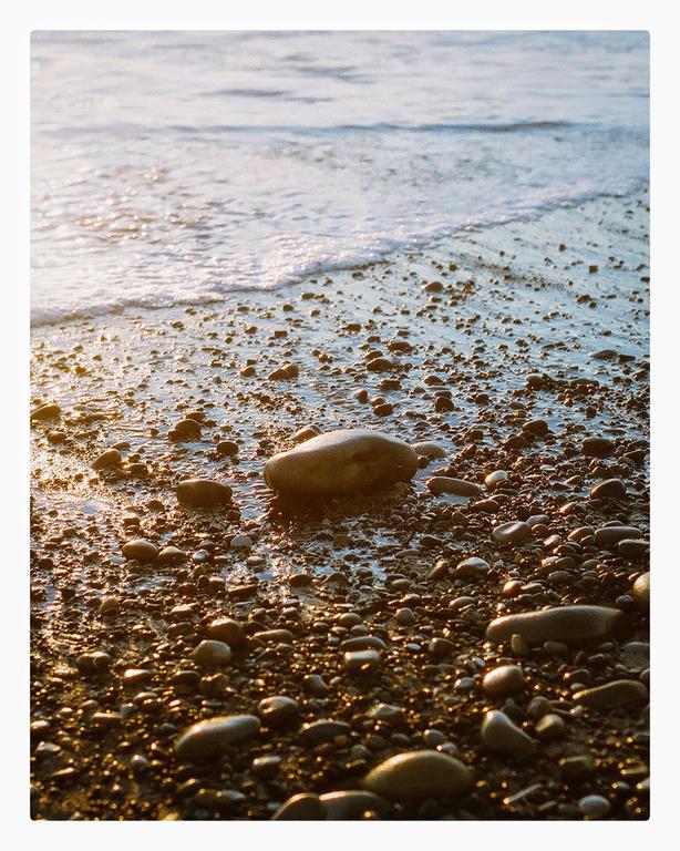 A close-up view shows wet rocks and pebbles on a beach with a gentle wave receding into the ocean.
