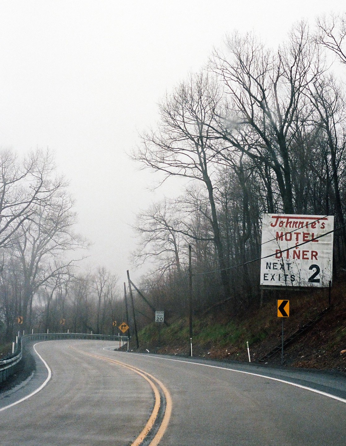 A winding road with a motel sign on the right is surrounded by bare trees under a hazy sky.