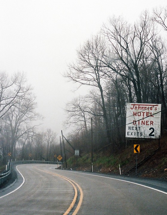 A winding road with a motel sign on the right is surrounded by bare trees under a hazy sky.
