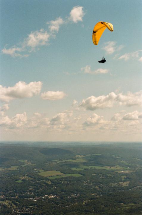 A paraglider with a yellow parachute flies high above a vast green landscape under a cloudy sky.