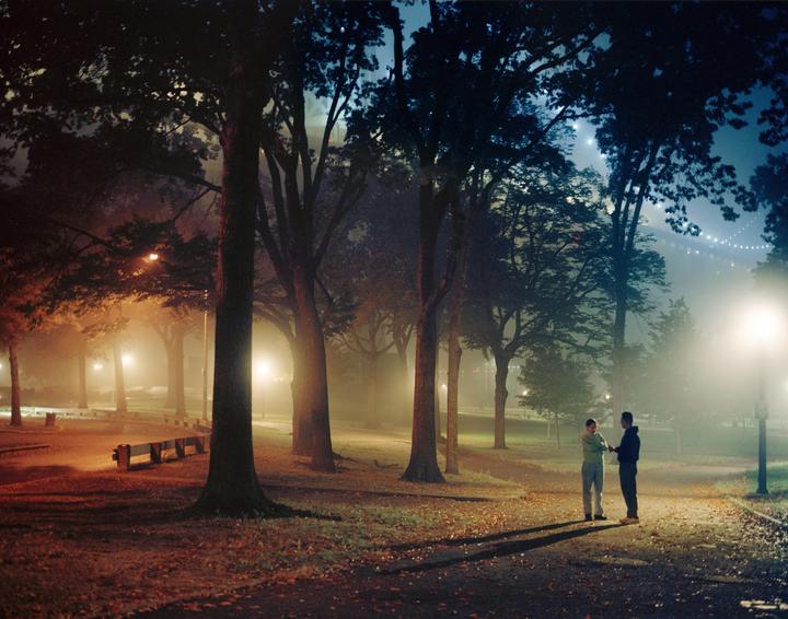 Two men stand on a path in a park at night, illuminated by streetlights and surrounded by trees and fog.