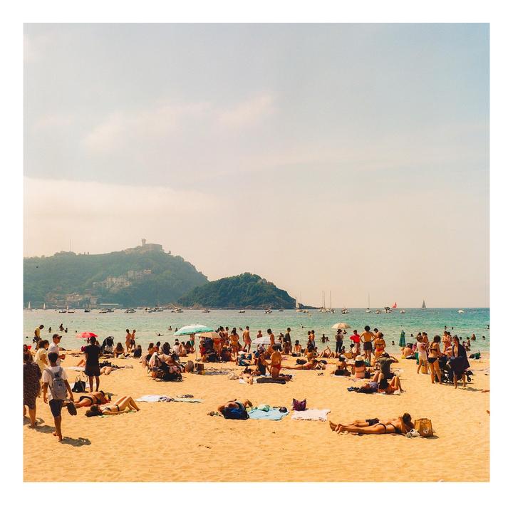 A crowded beach with people sunbathing and swimming, with mountains and boats in the background.