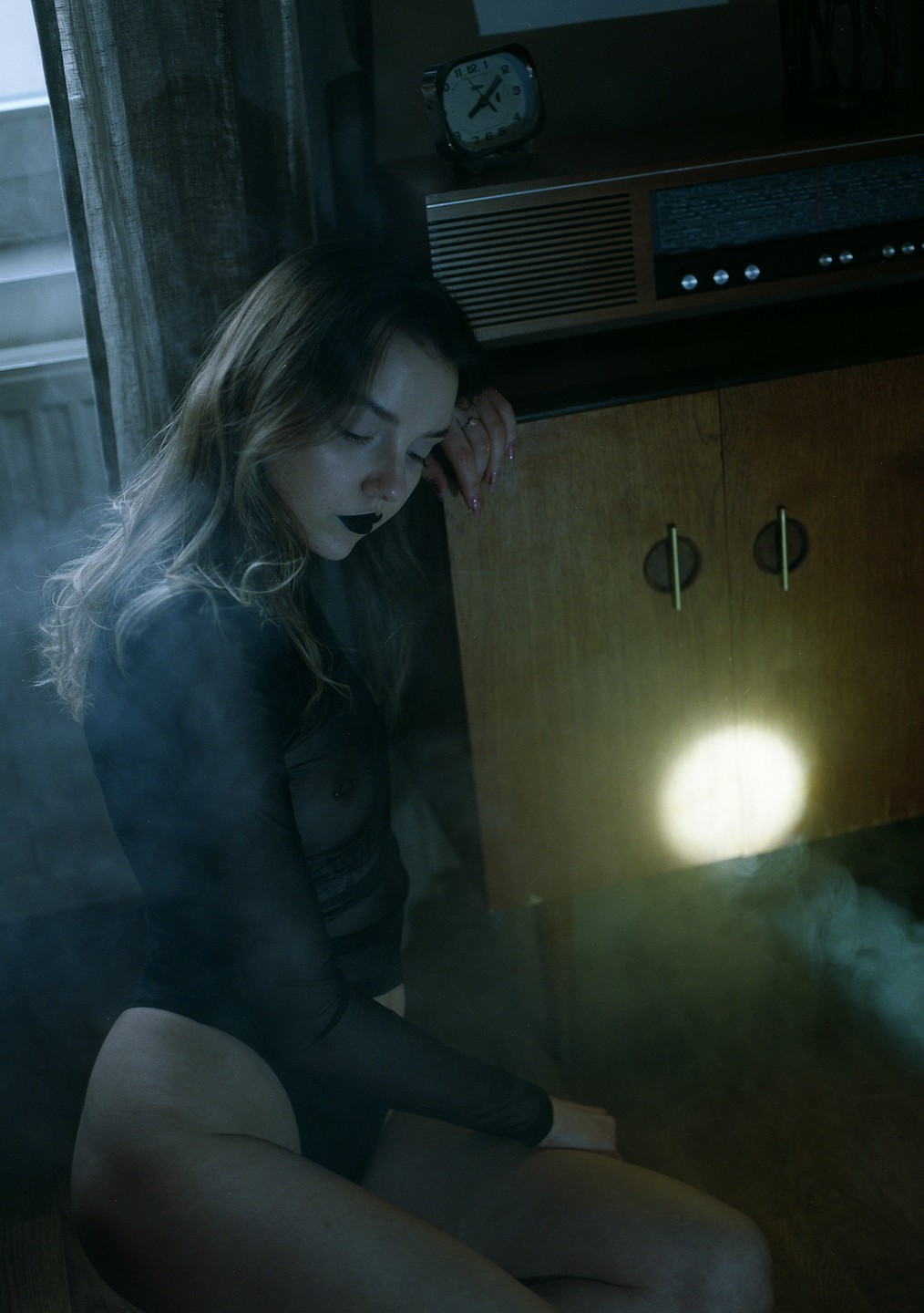 A woman with dark lipstick and a sheer top leans against a wooden cabinet with a radio and an alarm clock on top.