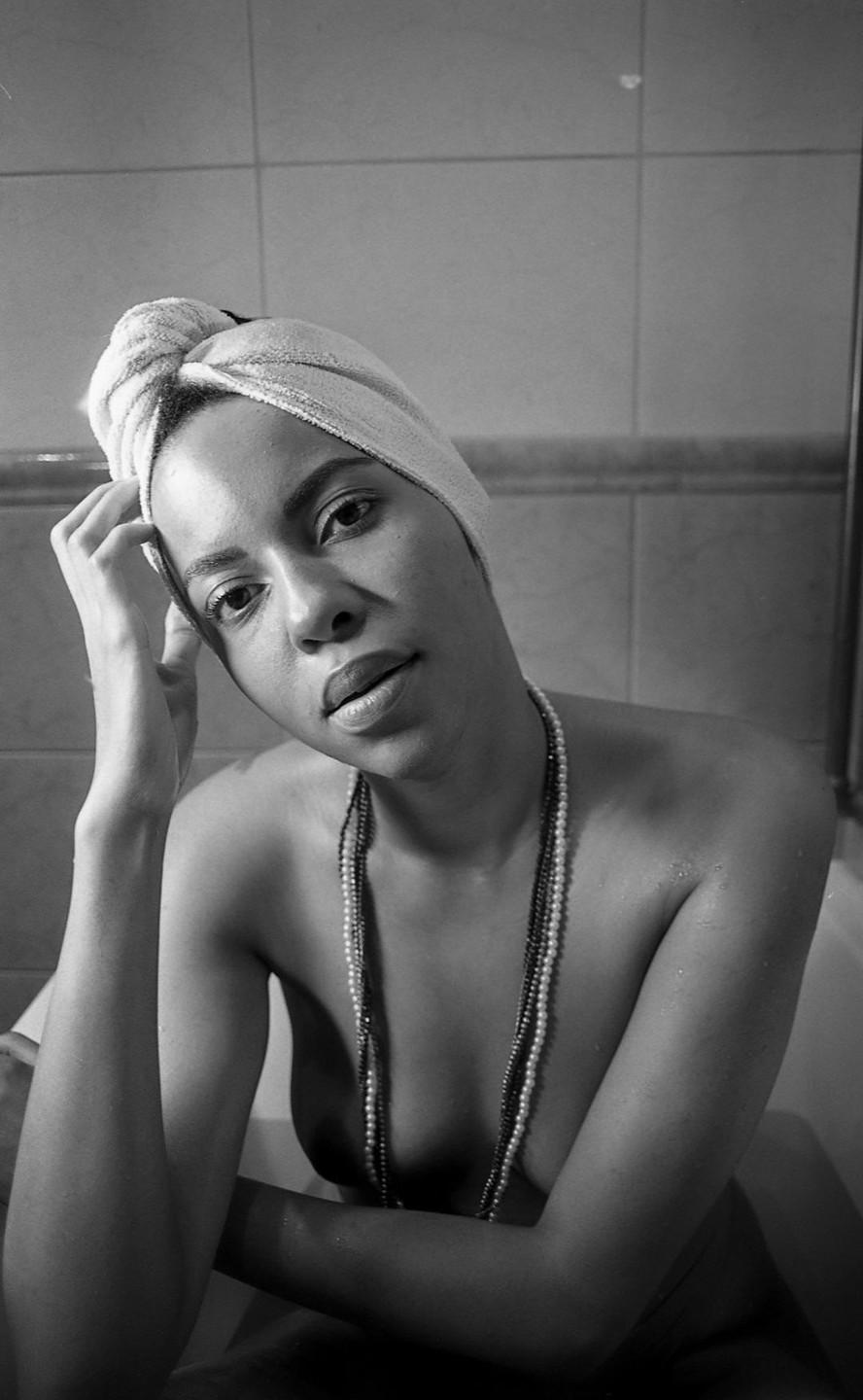 A woman wearing a towel on her head and multiple strands of beads sits in a bathtub.