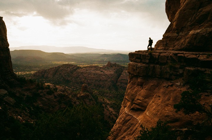 A person stands on a cliff overlooking a vast landscape of mountains and valleys under a cloudy sky.