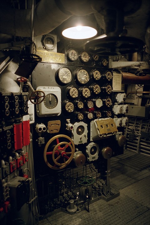 A control panel with many gauges, dials, and wheels is illuminated by an overhead light in a machine room.