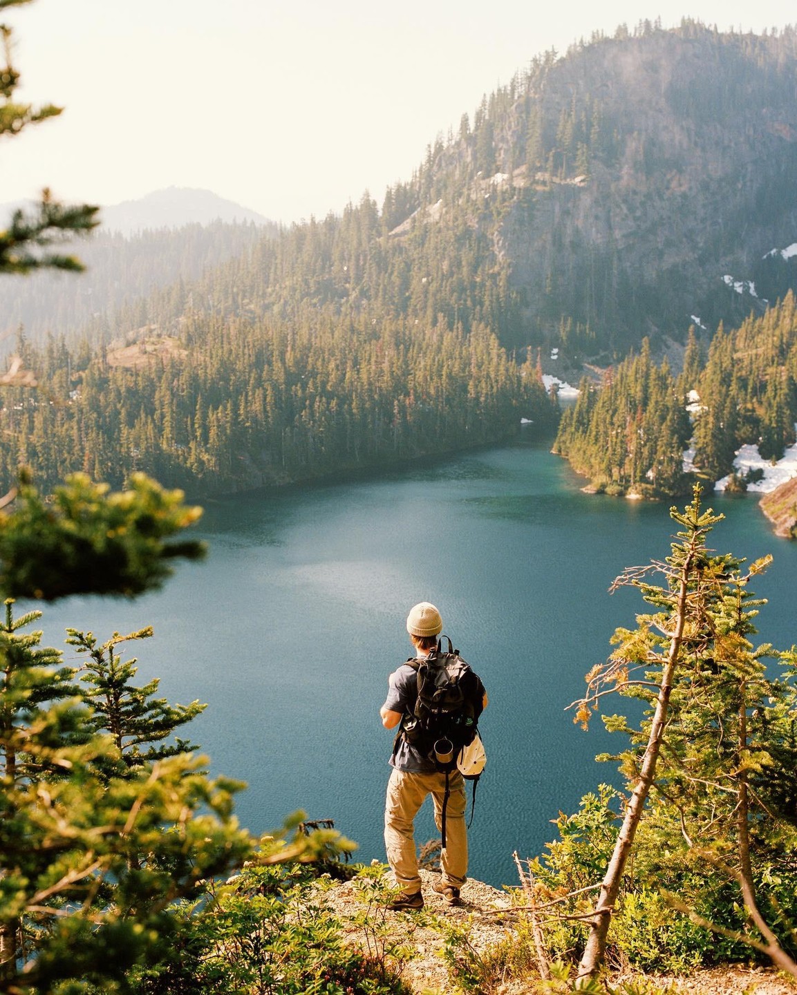 A man with a backpack stands on a cliff overlooking a lake surrounded by a forest.