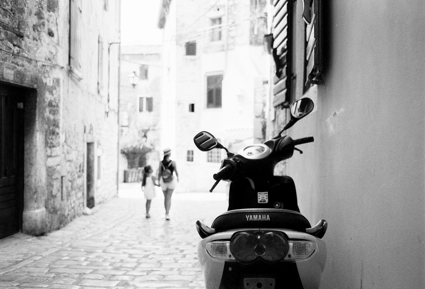 A scooter is parked on a narrow, cobbled street with a woman and child walking in the background.