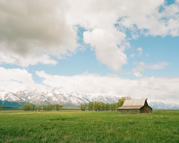 A wooden barn stands in a green field with snow-capped mountains and a cloudy sky in the background.