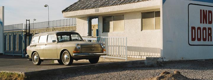 A beige station wagon is parked in front of a white building with a sign that reads "INDOOR" and a metal fence.