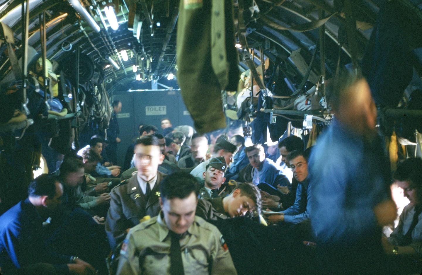 A group of men in military uniform are seated inside a dimly lit aircraft cabin with a "TOILET" sign visible in the background.