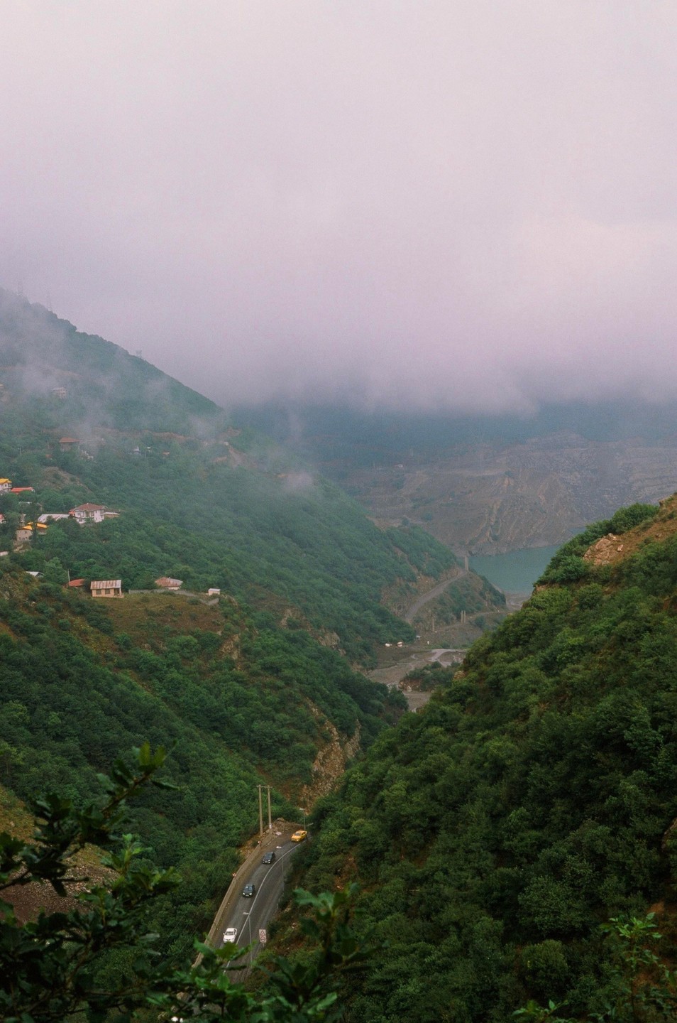 A winding road with cars and houses is visible on a lush green mountainside under a hazy sky.