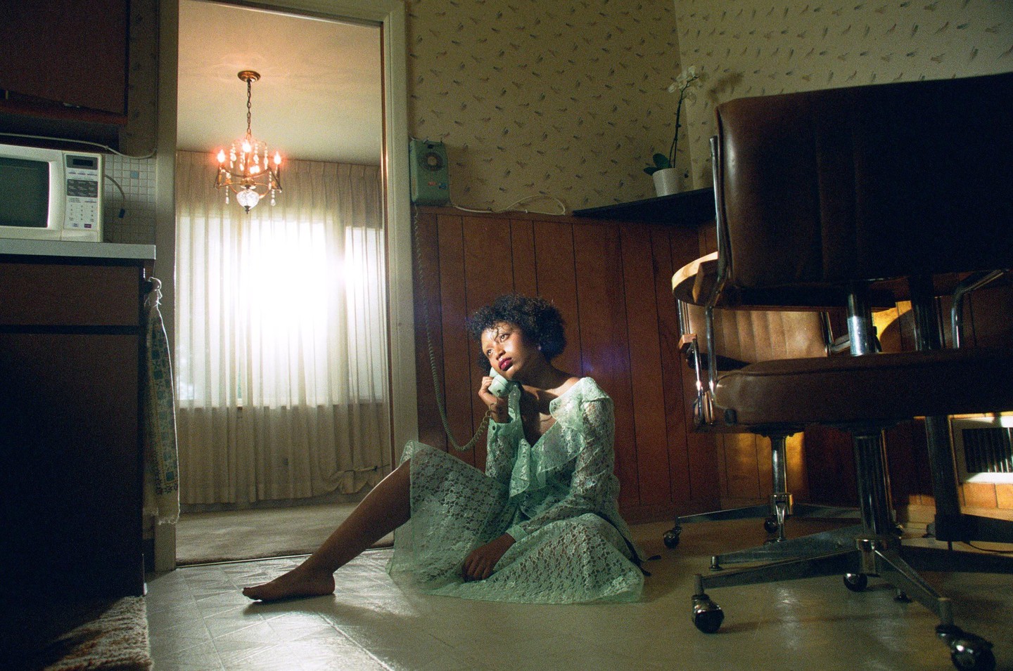 A woman in a lace dress sits on the floor and talks on a rotary phone in a room with wood paneling and patterned wallpaper.