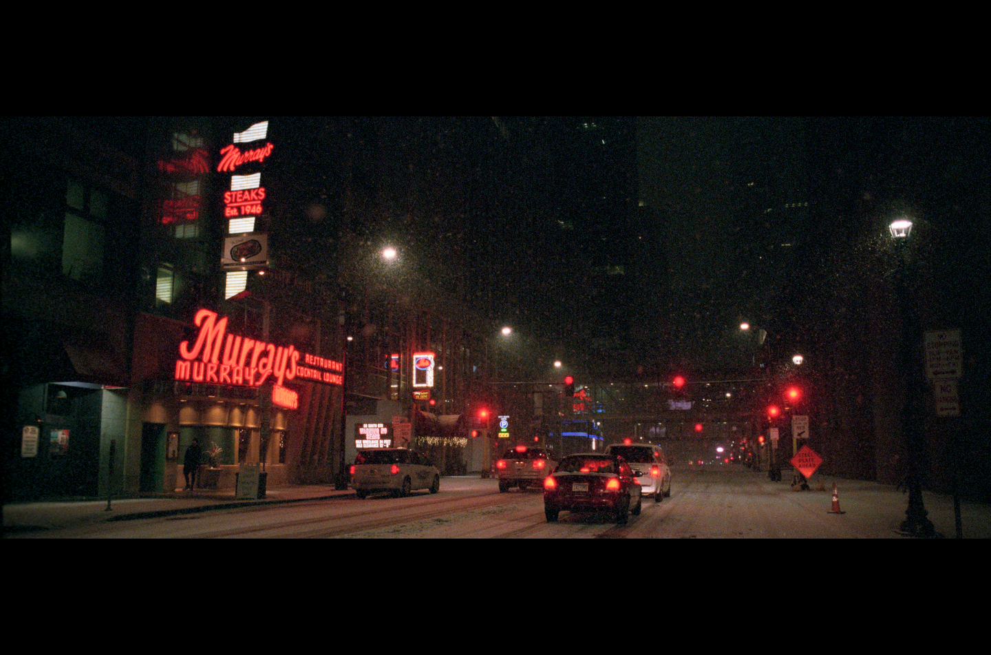 A snowy street at night is illuminated by neon signs and streetlights, with cars driving on the road and a restaurant on the left.