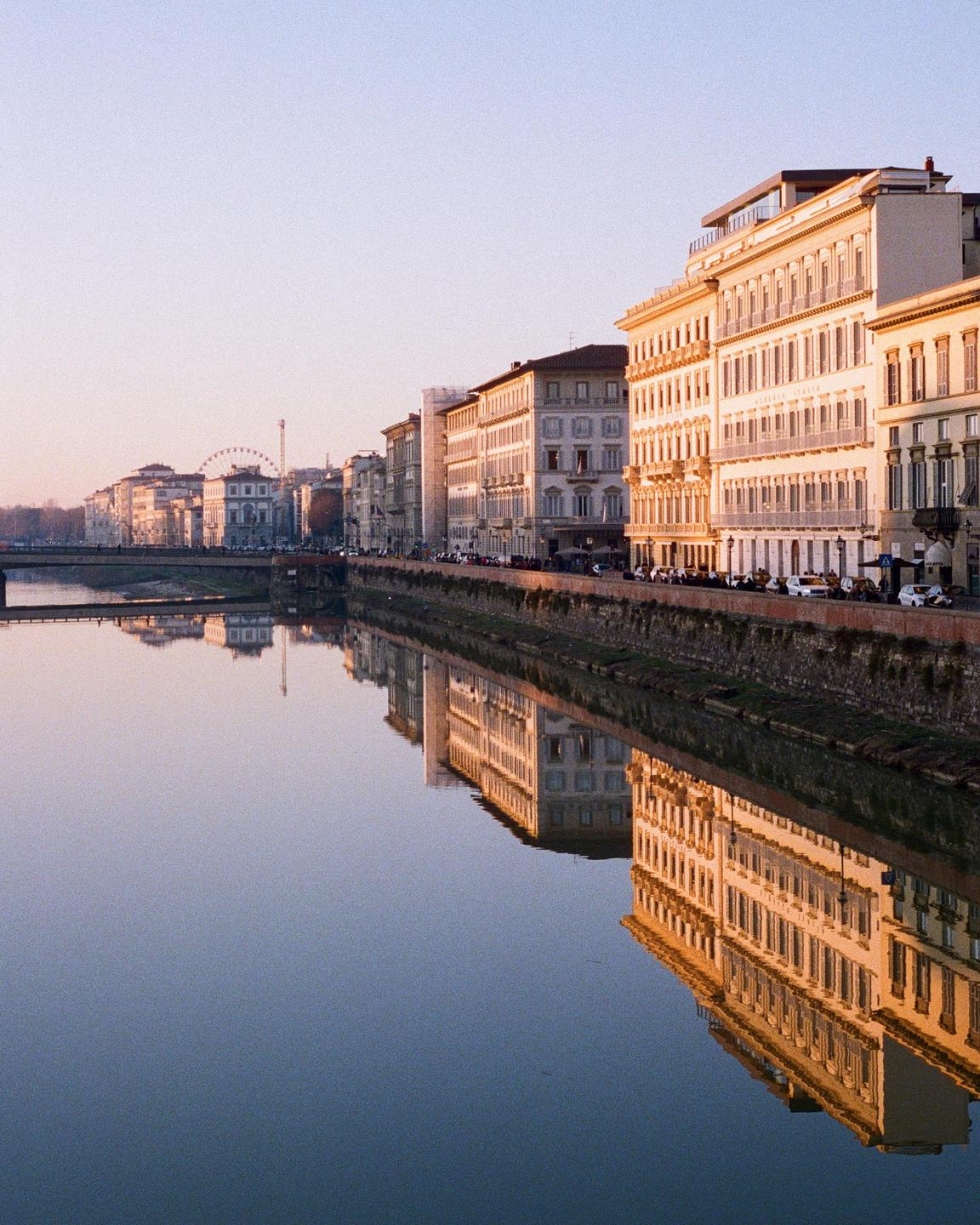A river reflects buildings and a bridge along its bank in Florence.