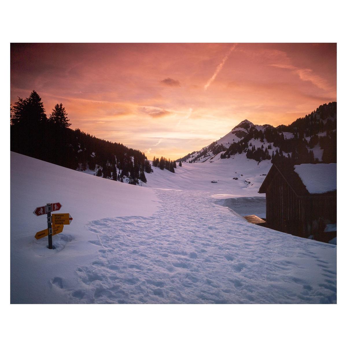 A snow-covered mountain valley with a wooden hut and a signpost is illuminated by the warm glow of sunset.