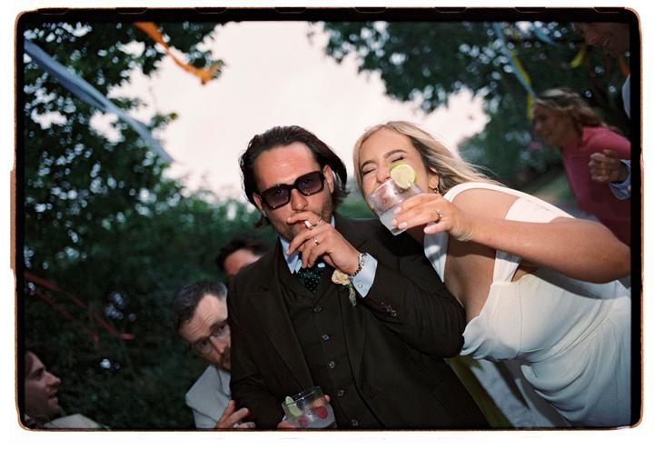 A man in sunglasses smokes a cigarette while a woman next to him laughs and holds a drink with a lime.