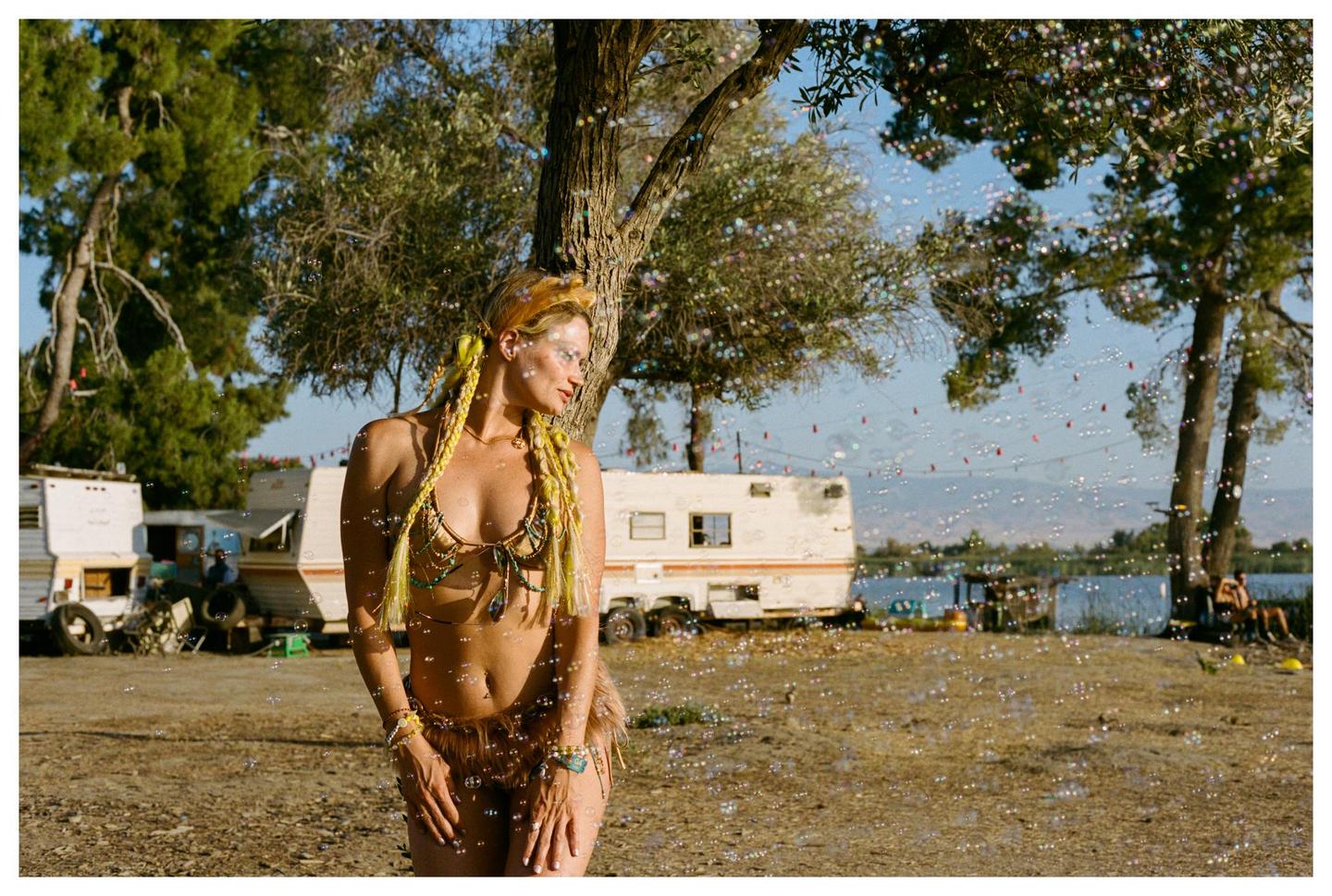 A woman with braided yellow hair and a beaded bikini stands outdoors with bubbles floating around her, with trailers and a body of water in the background.