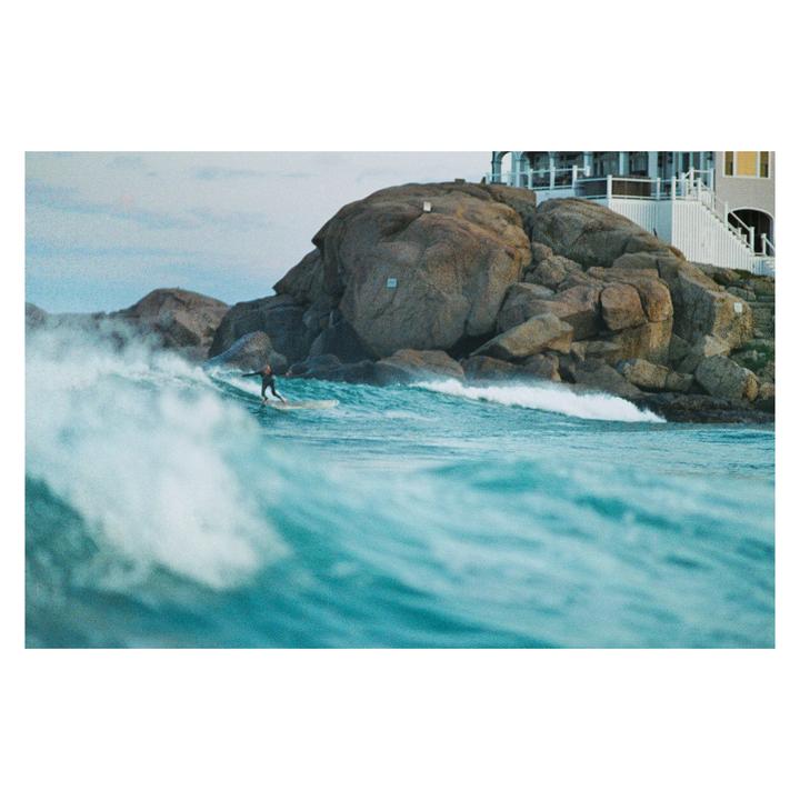 A surfer rides a wave in front of a rocky shore with a house on top.