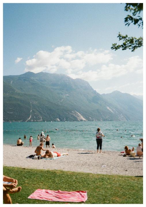 A group of people are swimming and relaxing on a beach next to a large lake with mountains in the background.