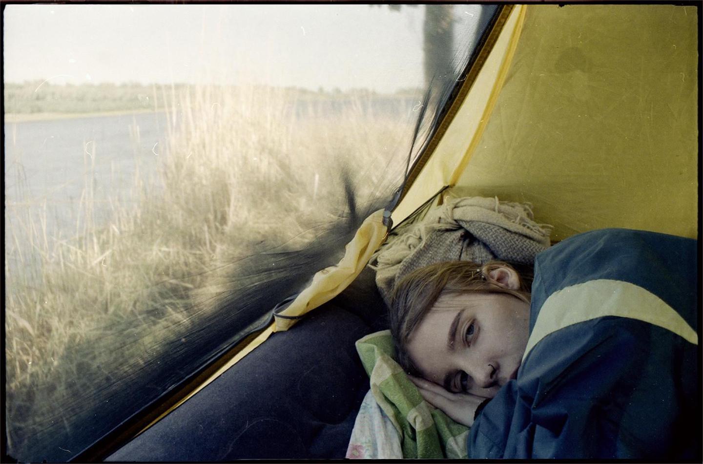A woman lies inside a tent looking out at a body of water and tall grass.