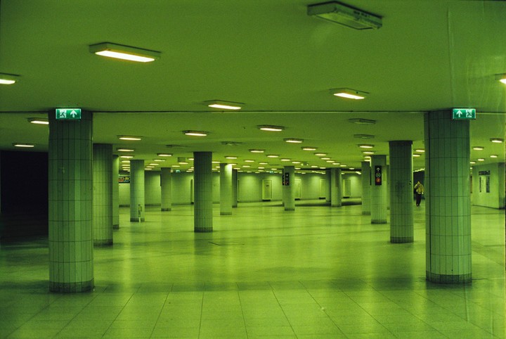 A person walks through a large, empty, green-lit underground hall with many pillars.