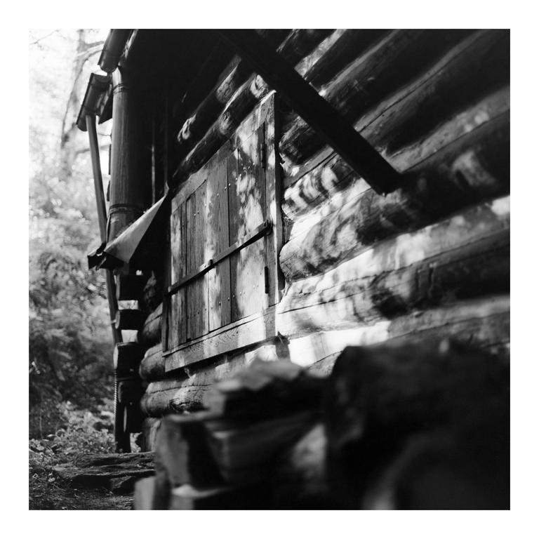 Sunlight casts shadows on the side of a rustic log cabin with a closed wooden window.