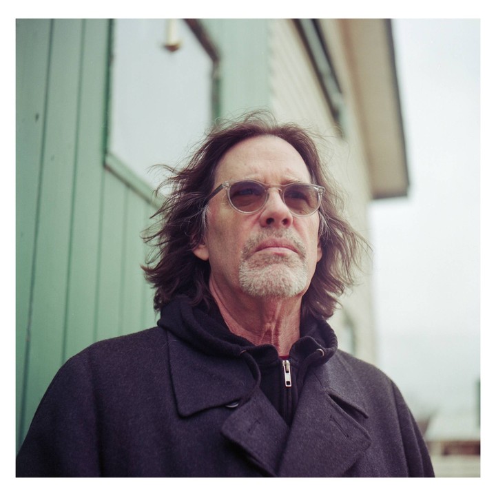 A man with long hair and a beard wears sunglasses and a dark coat while looking up.