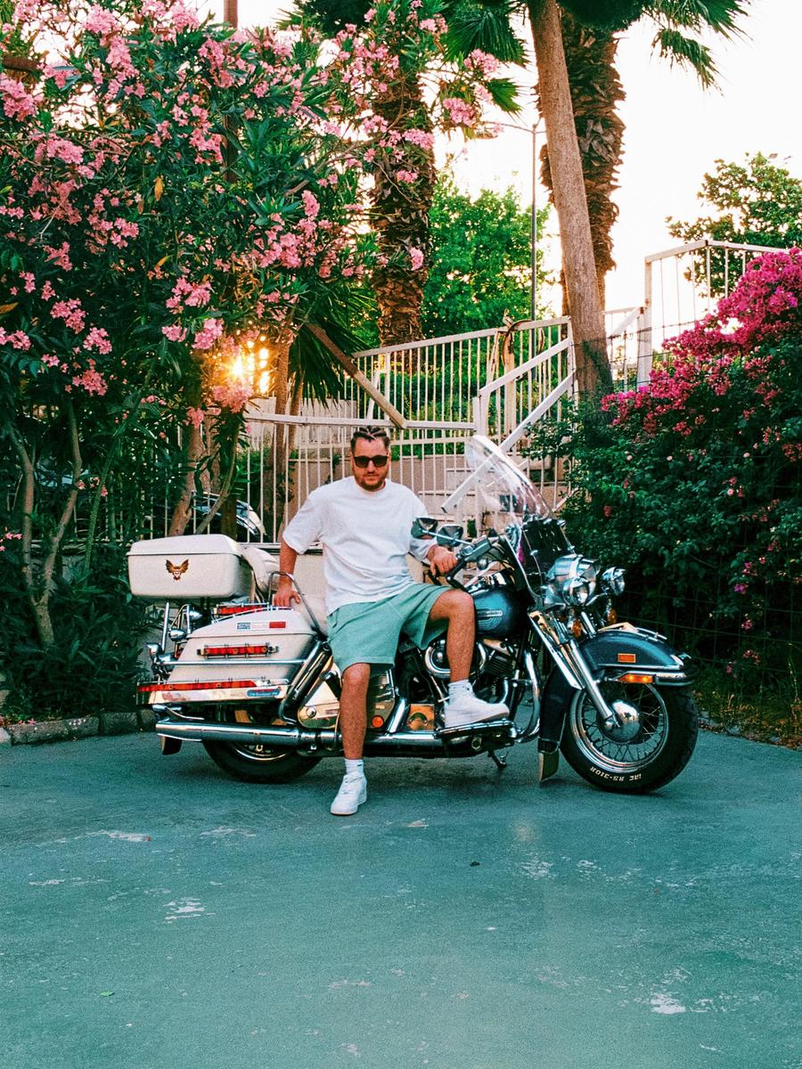 A man sits on a motorcycle surrounded by flowering bushes and palm trees.