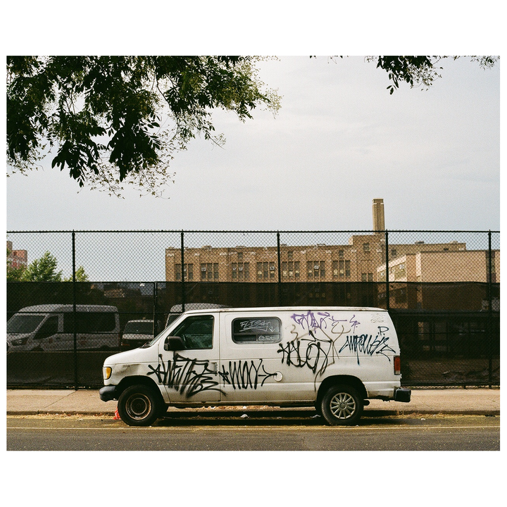 A white van covered in graffiti is parked on a street next to a chain link fence with a building in the background.