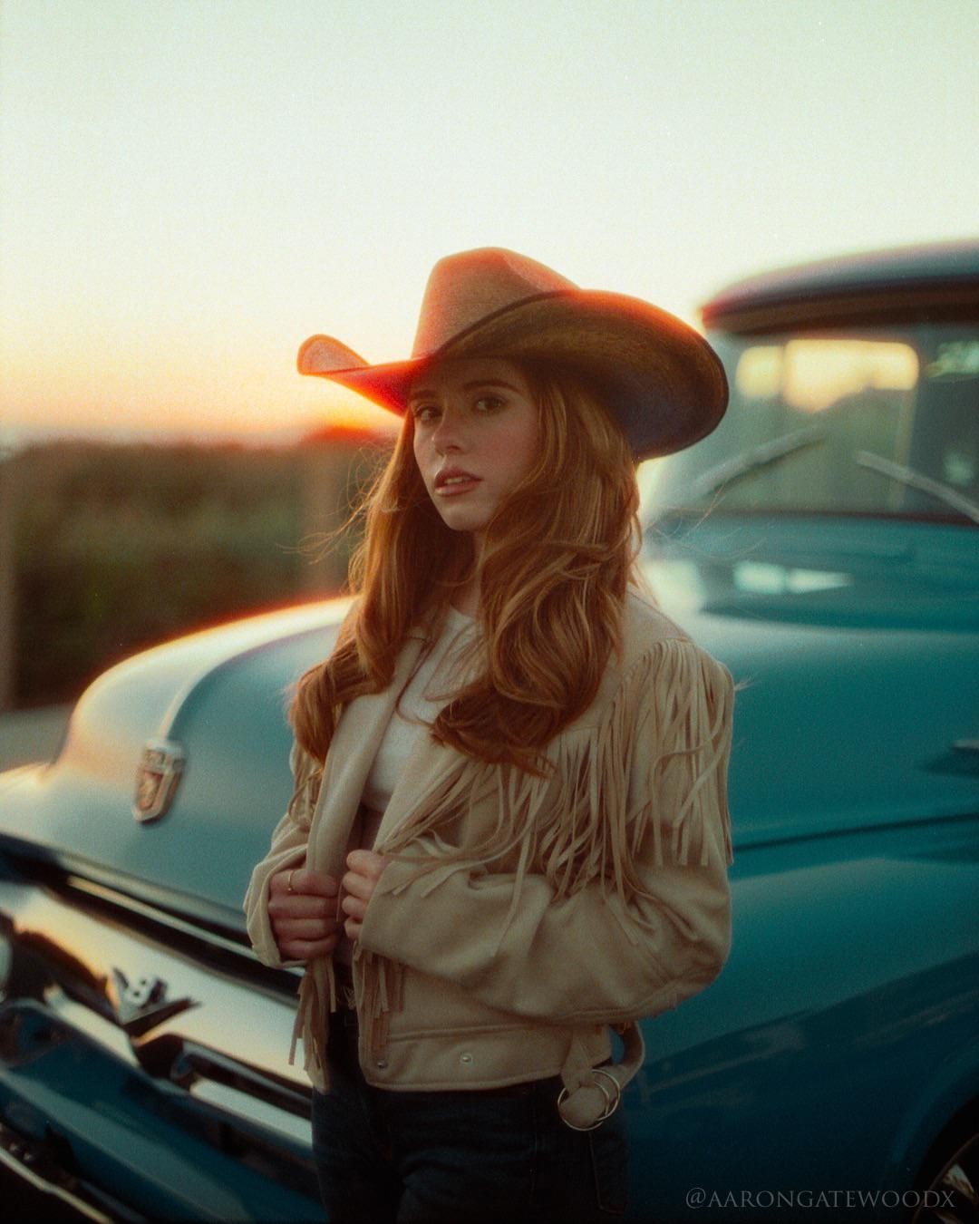 A woman in a cowboy hat and fringed jacket stands in front of a blue truck at sunset.