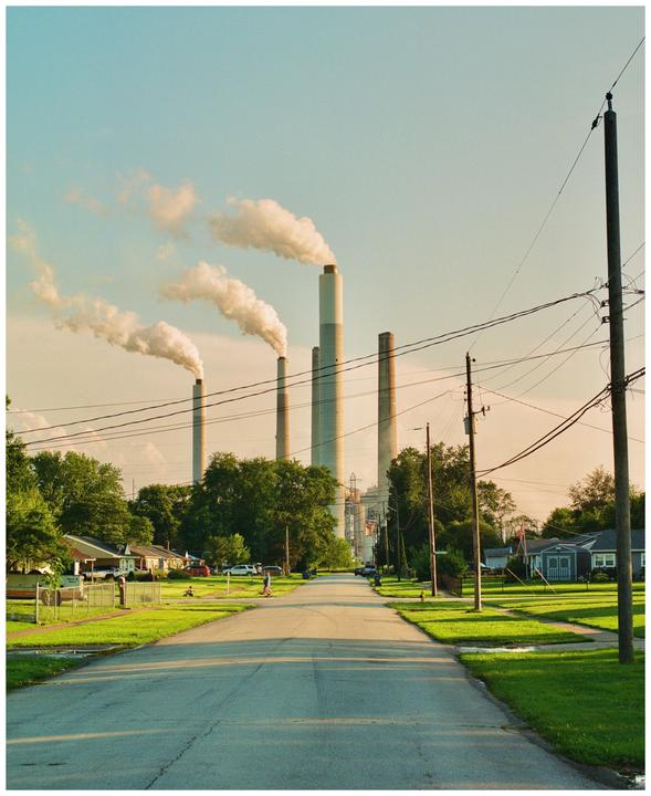 A street leads towards a power plant with multiple smoking smokestacks in the distance.