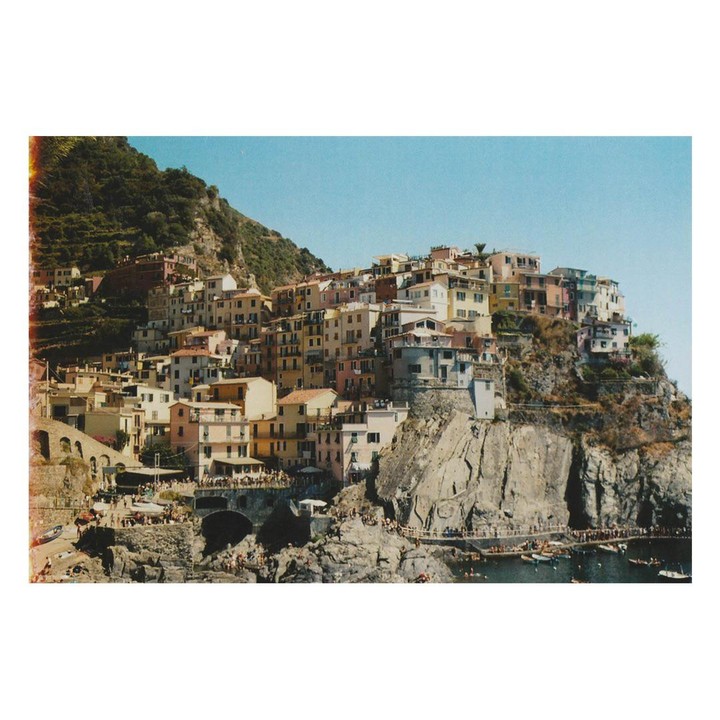 A colorful village is built into a steep hillside overlooking the sea, with many people on the rocks and in the water below.