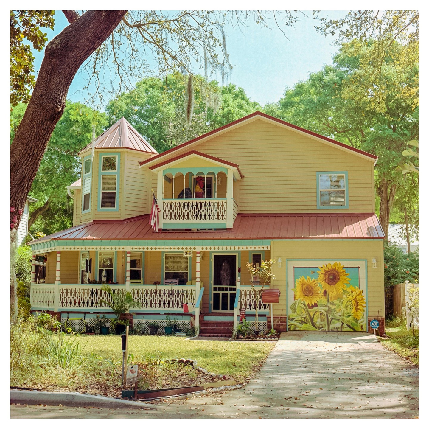 A yellow house with a red roof and a sunflower mural on the garage door stands among green trees.