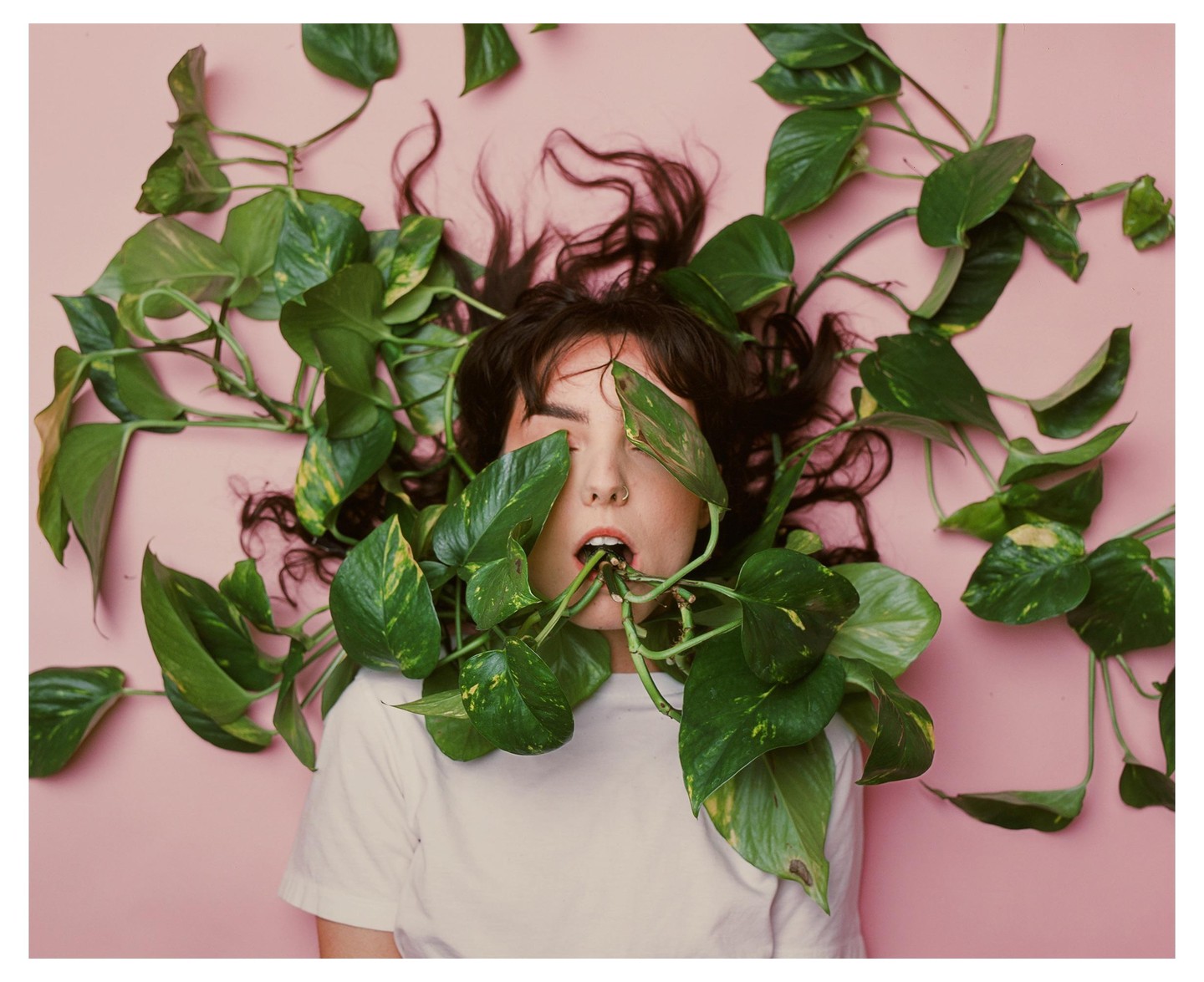 A woman lies on a pink surface with green plants covering her face and body.