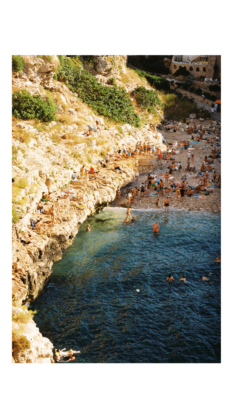 A rocky cove is filled with people swimming in the clear blue water and sunbathing on the shore.