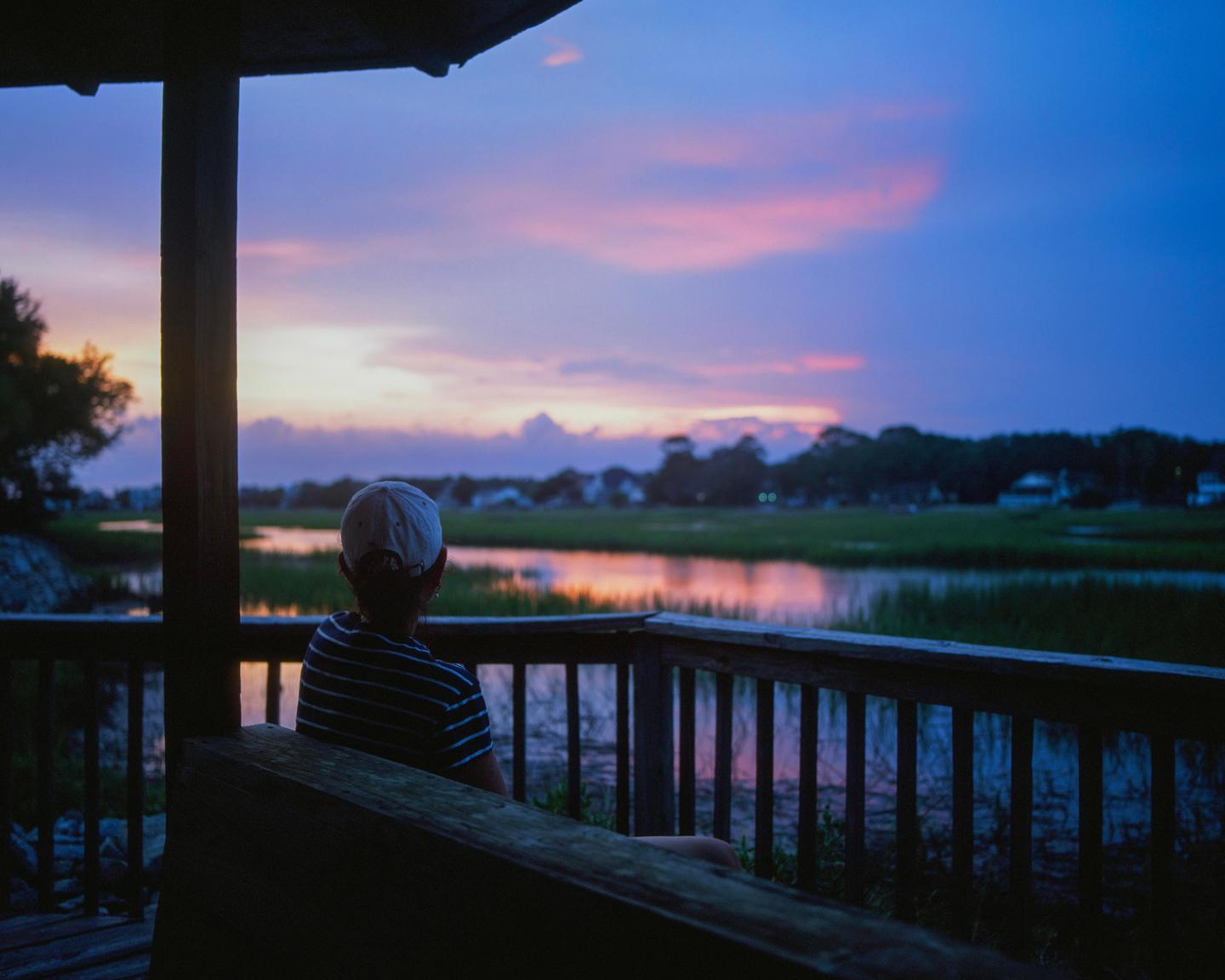 A person wearing a baseball cap sits on a porch overlooking a body of water with a colorful sunset.