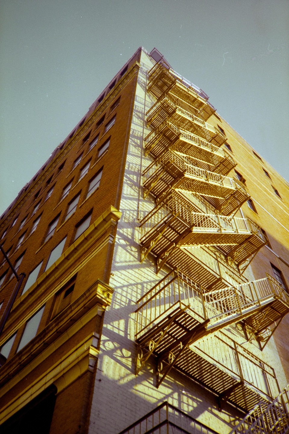 A low-angle view shows a tall brick building with many windows and a metal fire escape on its side, casting shadows on the wall.