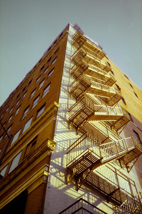 A low-angle view shows a tall brick building with many windows and a metal fire escape on its side, casting shadows on the wall.