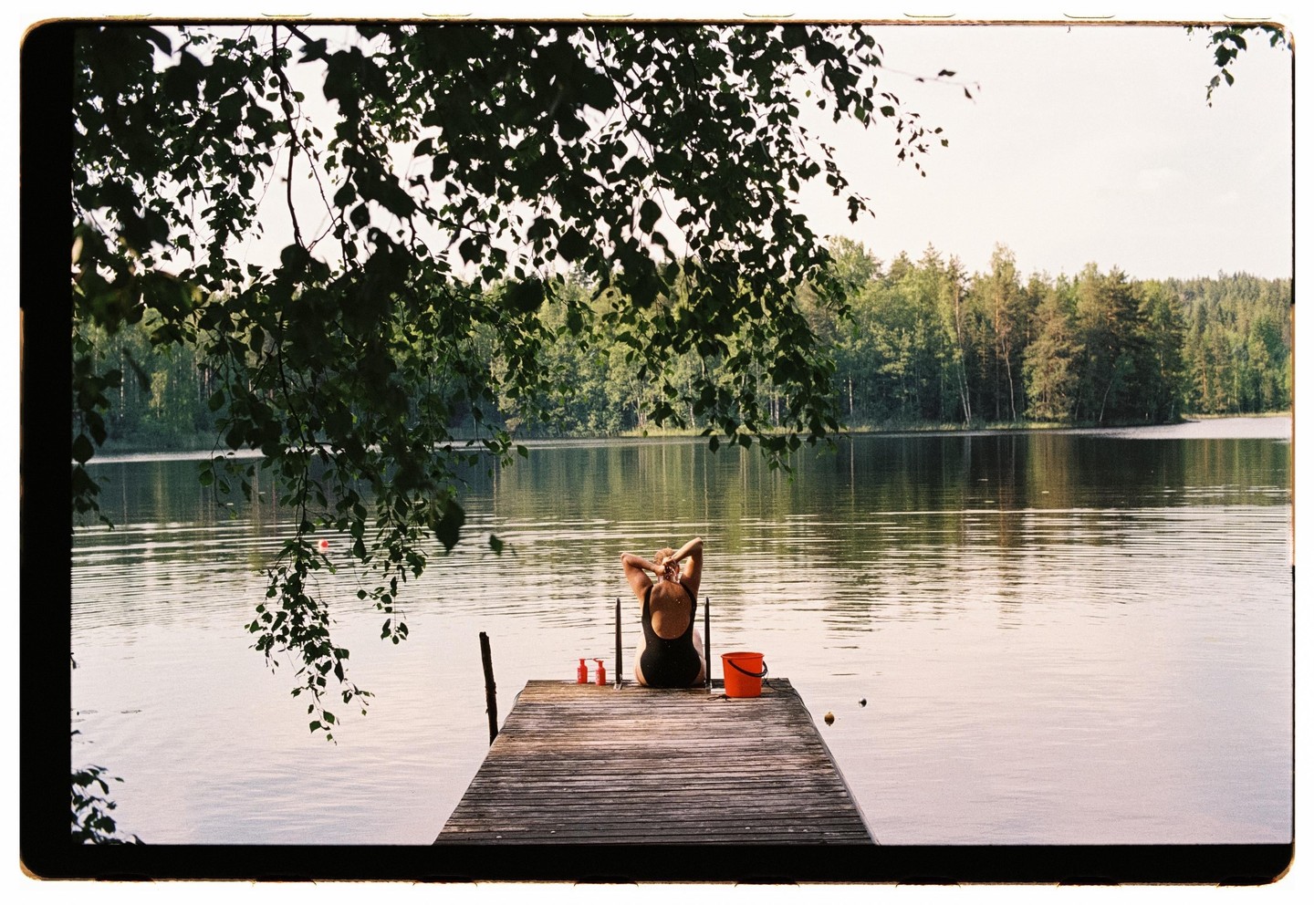 A woman sits on a wooden dock by a lake, with trees on the far bank and leaves hanging over the water in the foreground.