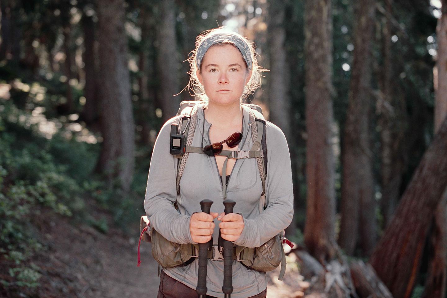 A woman wearing a backpack and holding trekking poles stands on a trail in a forest.