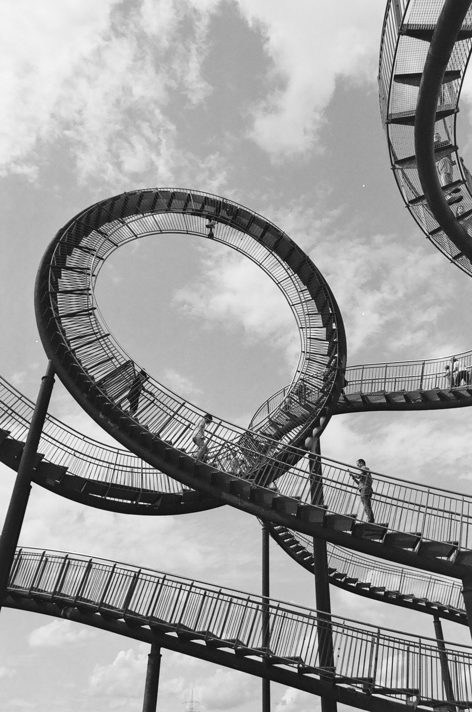People walk on a large, looping metal sculpture with stairs against a cloudy sky.