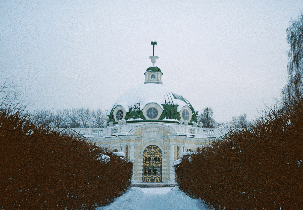 A snow-covered building with a green dome and ornate golden doors is framed by dark bushes and a snowy path.