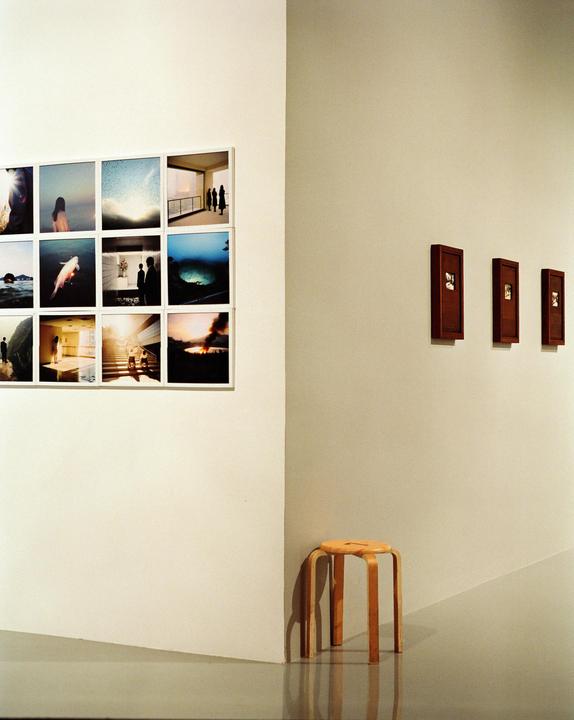 A wooden stool sits in the corner of an art gallery with several framed photographs on the walls.