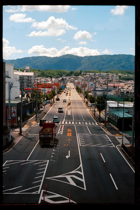 A wide street with cars and buildings on either side leads towards distant mountains under a blue sky with clouds.