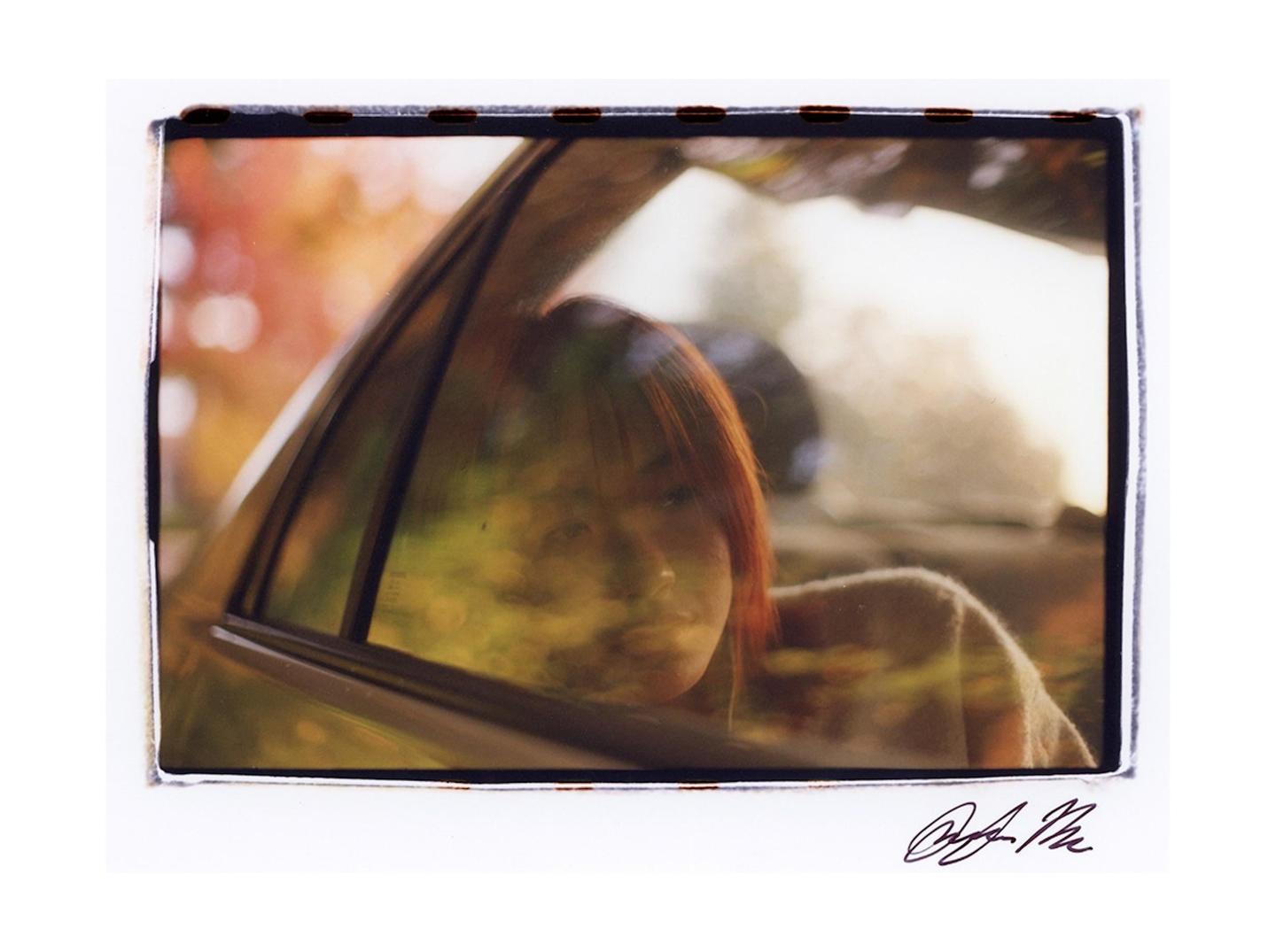A woman with red hair looks out of a car window, with reflections of autumn foliage visible on the glass.