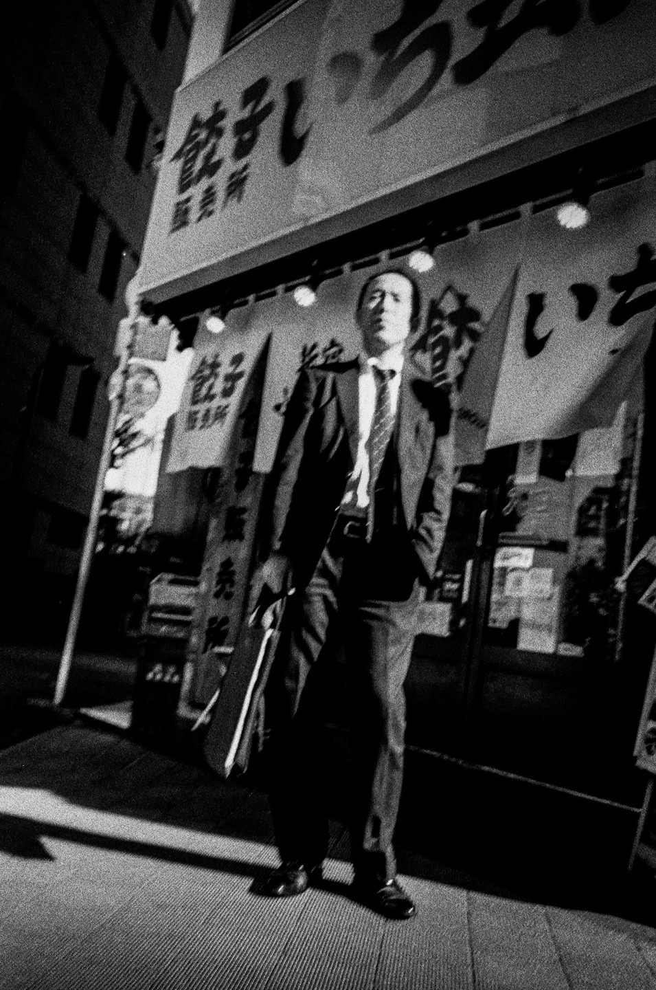 A man in a suit stands on a street at night with signs and banners in the background.