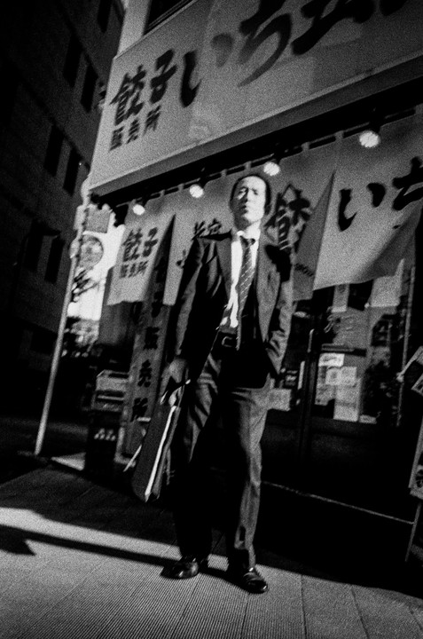 A man in a suit stands on a street at night with signs and banners in the background.