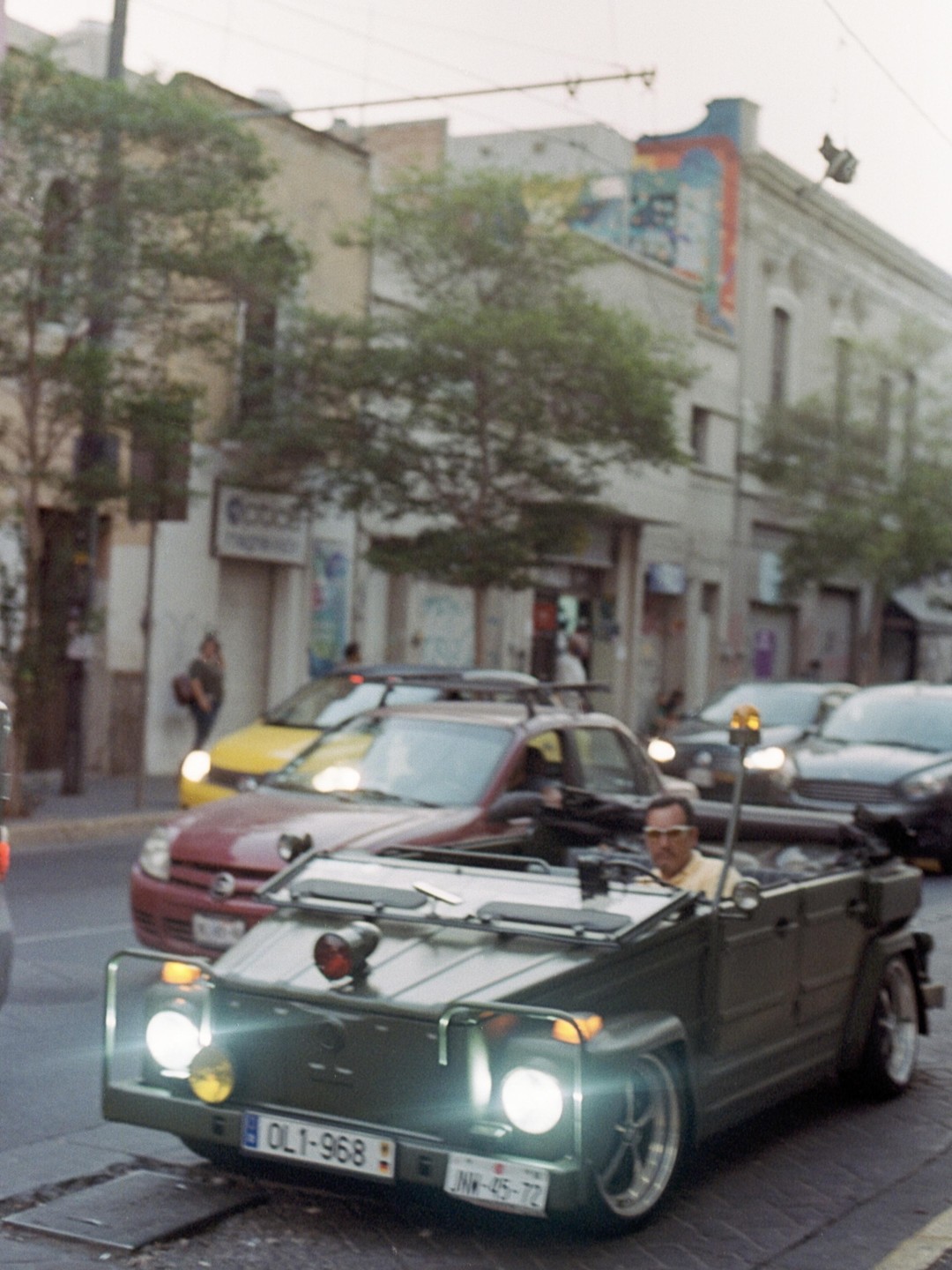 A man drives a green convertible car with its headlights on down a street in Guadalajara, Mexico.