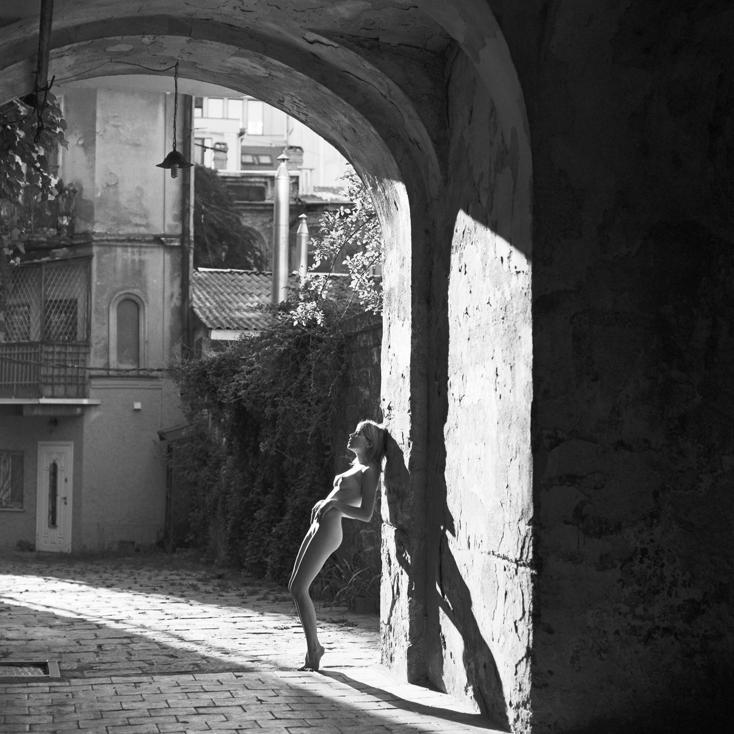 A nude woman leans against a sunlit wall under an archway in a cobblestone alley.